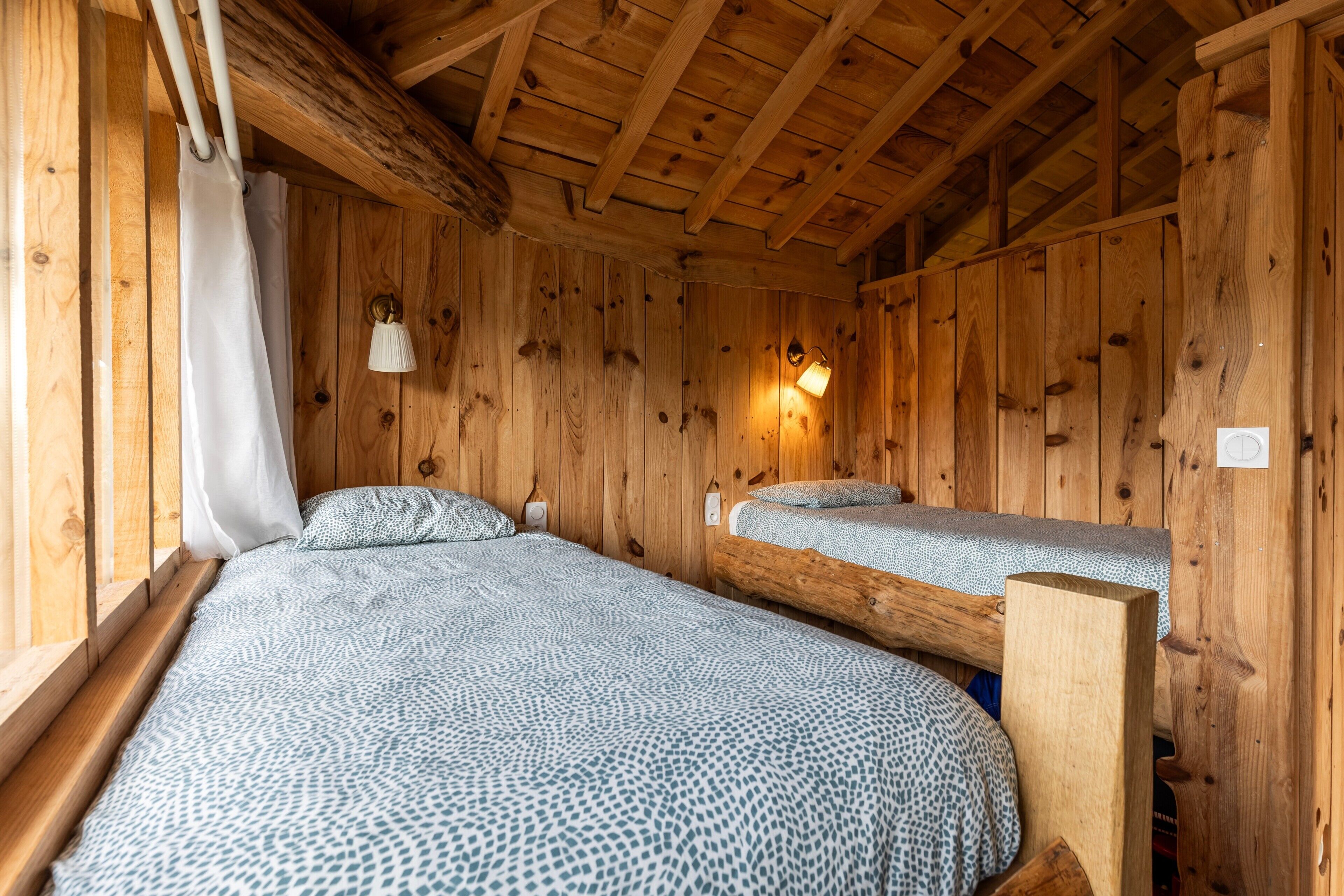 Una habitación tipo cabaña de madera con dos camas, una ventana con cortinas, una lámpara de pared y techo de madera.