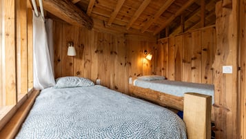Una habitación tipo cabaña de madera con dos camas, una ventana con cortinas, una lámpara de pared y techo de madera.