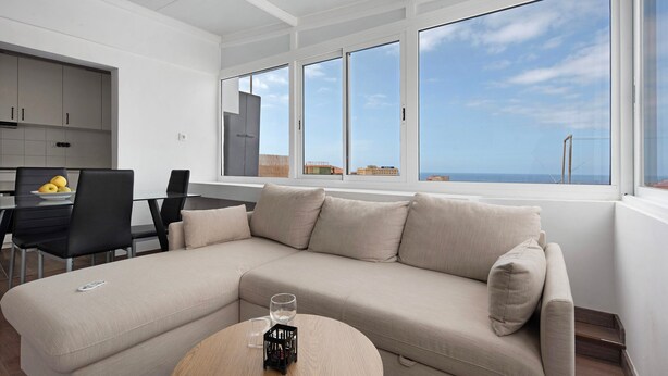 Una sala de estar moderna con un sofá color beige, una mesa redonda y un comedor con una mesa y sillas. Amplias ventanas ofrecen vista al mar.