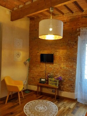 Une pièce aux murs en briques apparentes, avec un plafond en bois, un tapis rond en fibres naturelles, une chaise jaune, une petite table sur laquelle est posé un vase, et un téléviseur fixé au mur.
