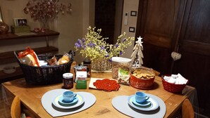 A wooden dining table with two blue placemats, a basket of bread, a jar of jam, a coffee pot, a container of biscotti, and a vase with flowers. - Casa Clery Tuscan Style Breakfast Included (Montelupo Fiorentino)