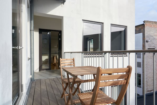 A wooden deck with a table and chairs, a white railing, and a building entrance.