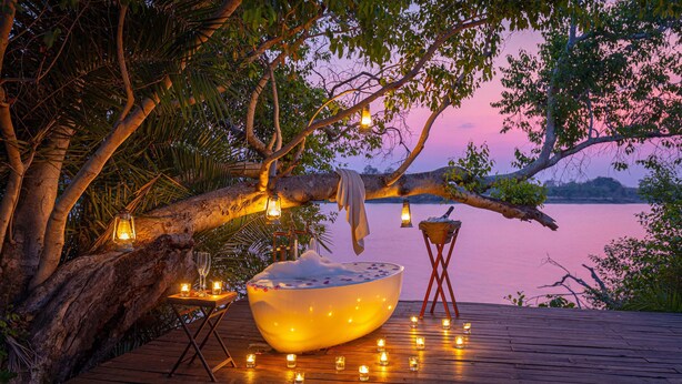 Une terrasse en bois avec un grand récipient, des bougies et des lanternes, le tout installé sous un arbre, près d’une étendue d’eau.