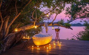 A wooden deck with a large bowl, candles, and lanterns under a tree by a body of water.