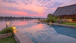 Un espace paisible au bord du lac, avec une piscine et une structure à toit de chaume, entouré d’une végétation luxuriante et illuminé le soir.