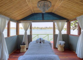 Una habitación con techo de madera, una cama con manta blanca, una araña y vista a un lago.