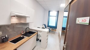 Una cocina compacta con armarios blancos, un lavabo negro y una encimera de madera. Hay una ventana con cortinas azules y una puerta con un cartel que dice "3D".