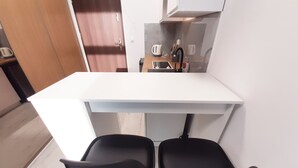 Una pequeña kitchenette con una encimera blanca, dos sillas negras y una puerta de madera al fondo.