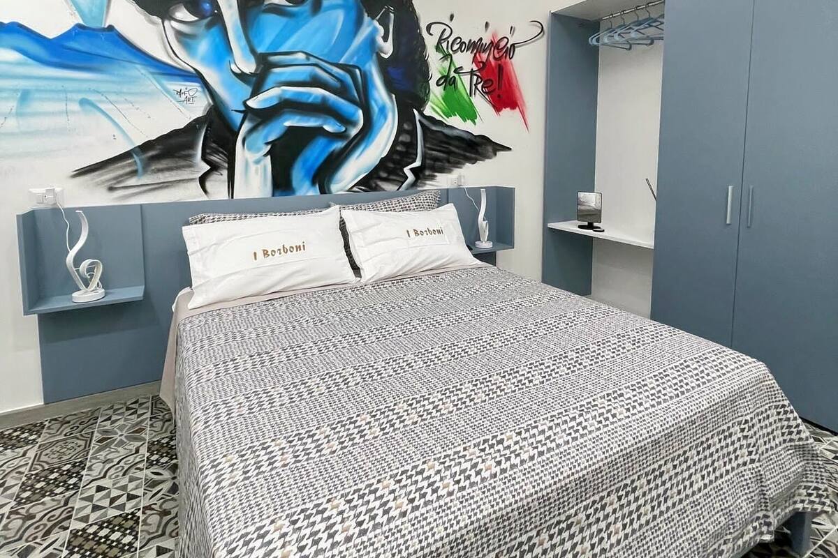 A bedroom with a large mural, a bed with patterned bedding, a wardrobe, and a mirror.