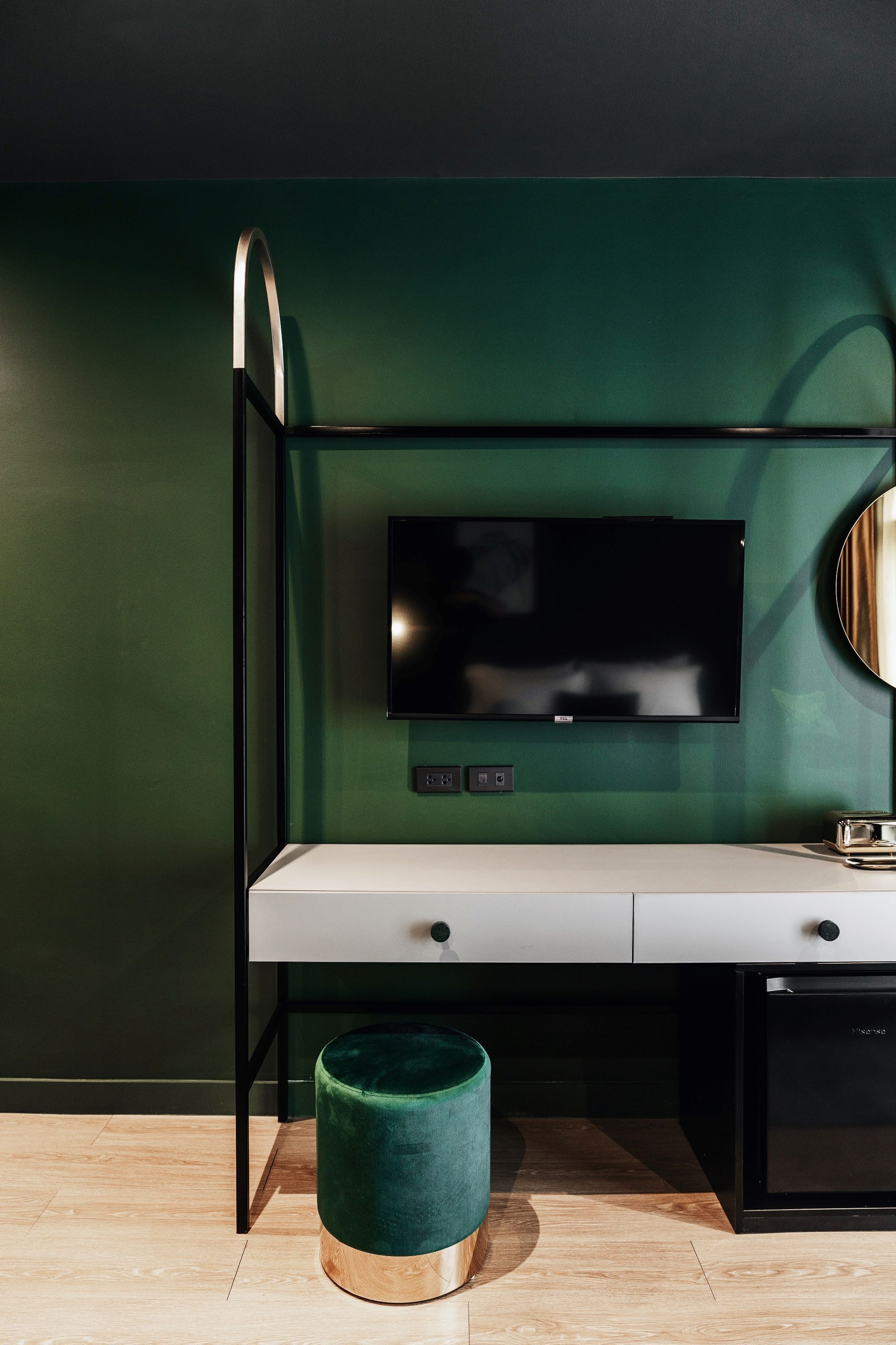 A modern room with a TV mounted on a green wall, a white desk, a black cabinet, and a green ottoman.