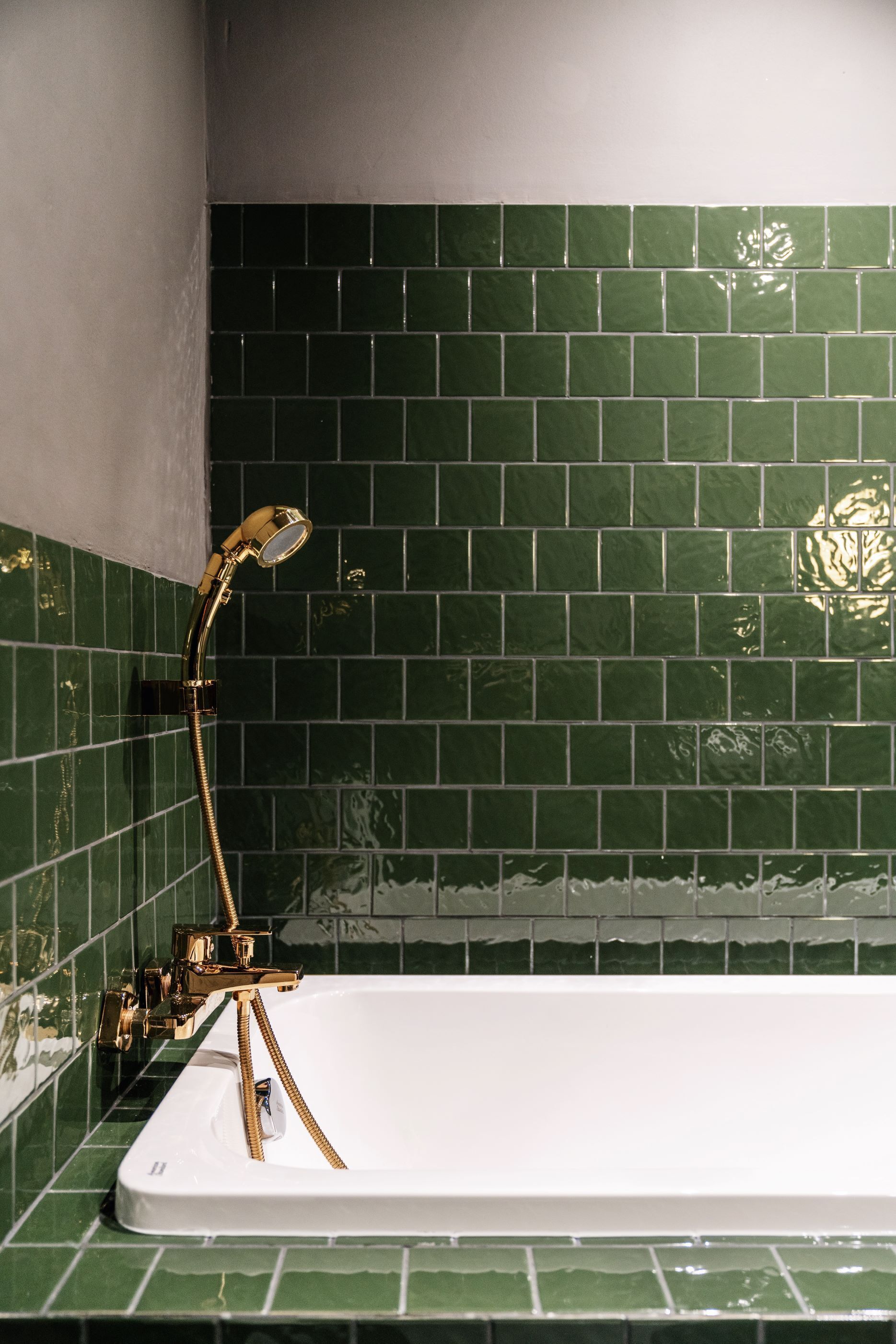 A bathtub with a gold faucet and hose, set against a green-tiled wall.