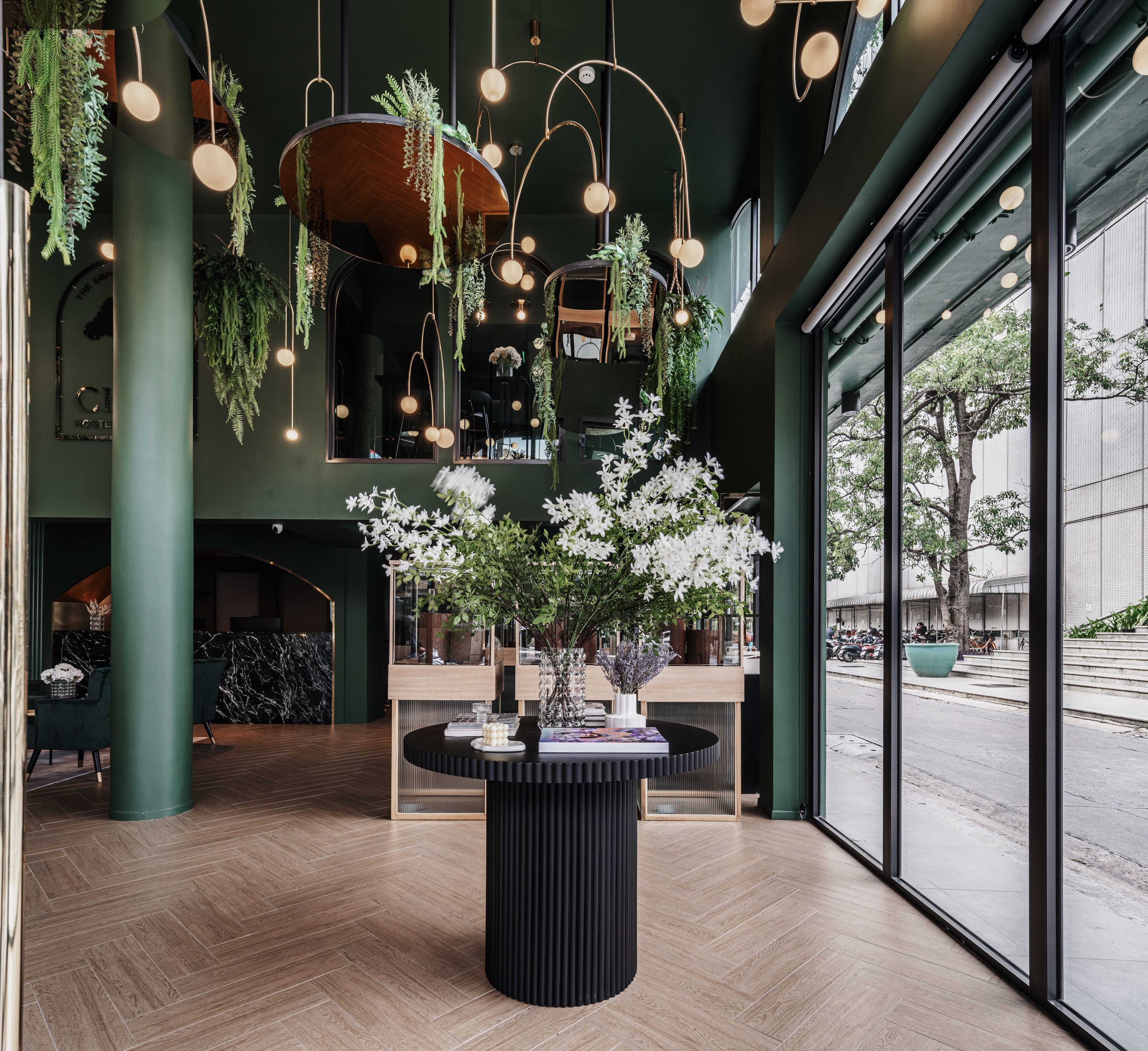 A modern interior with a central black table surrounded by books, a large window with a view of the street, and hanging plants.