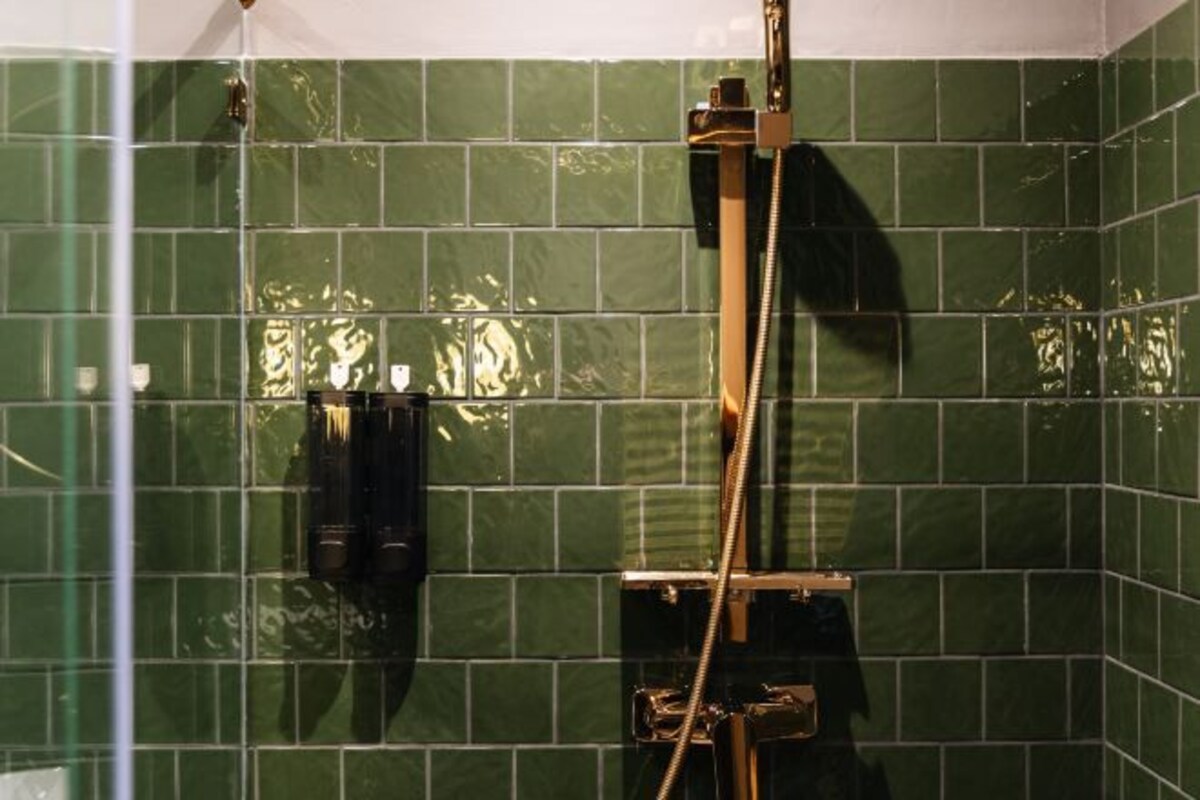 A bathroom with green tiled walls and a shower with a gold fixture.