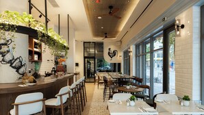 Ein Restaurant mit Barbereich, Esstischen und Stühlen. An Wänden und Decke befinden sich Pflanzen, und an der Wand ist ein Hahn-Mural zu sehen.