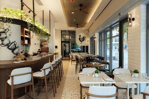 A restaurant with a bar area, dining tables, and chairs. There are plants on the walls and ceiling, and a rooster mural on the wall.