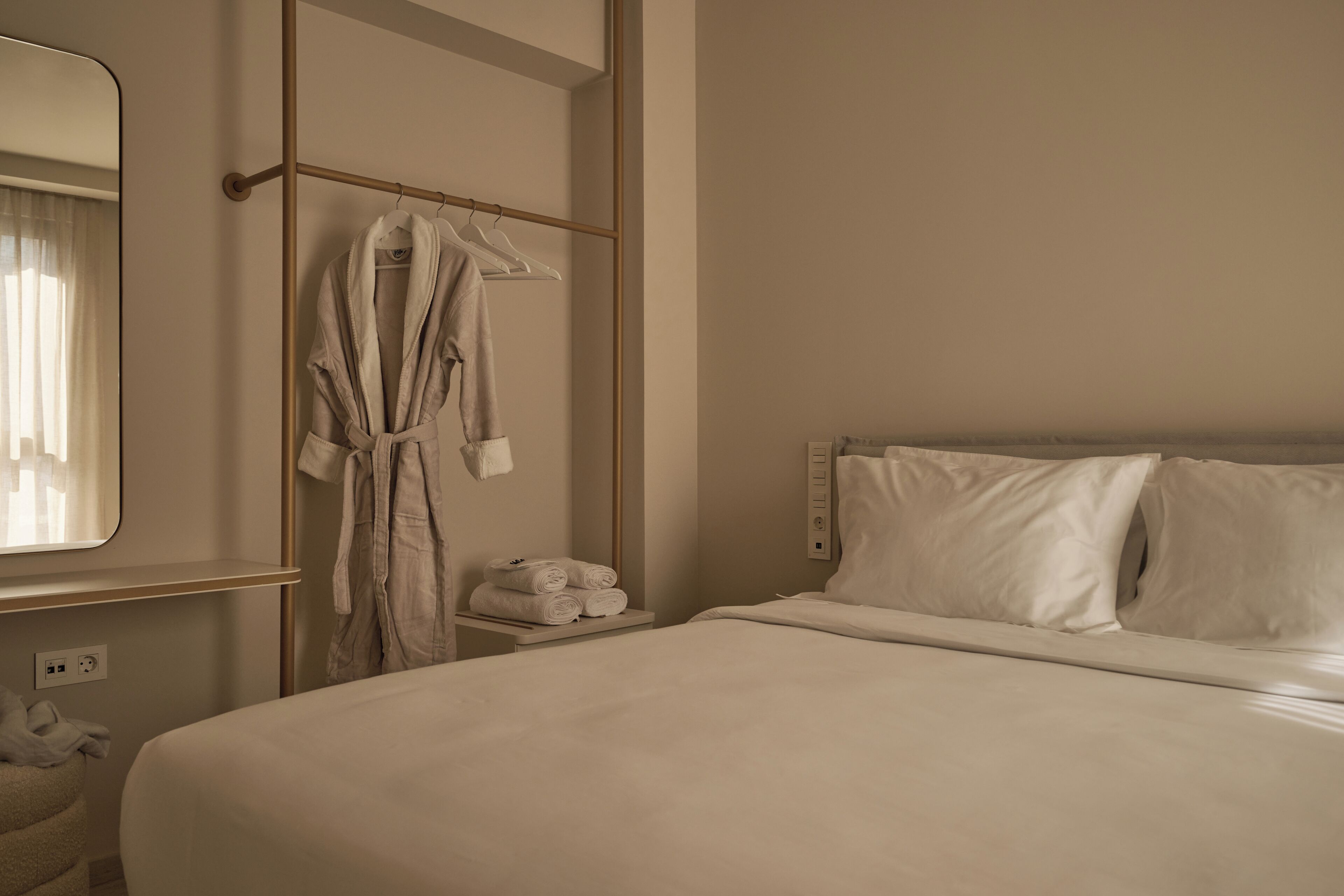 A hotel room with a large bed, a bedside table with a lamp, a robe hanging on a rack, and a mirror on the wall.