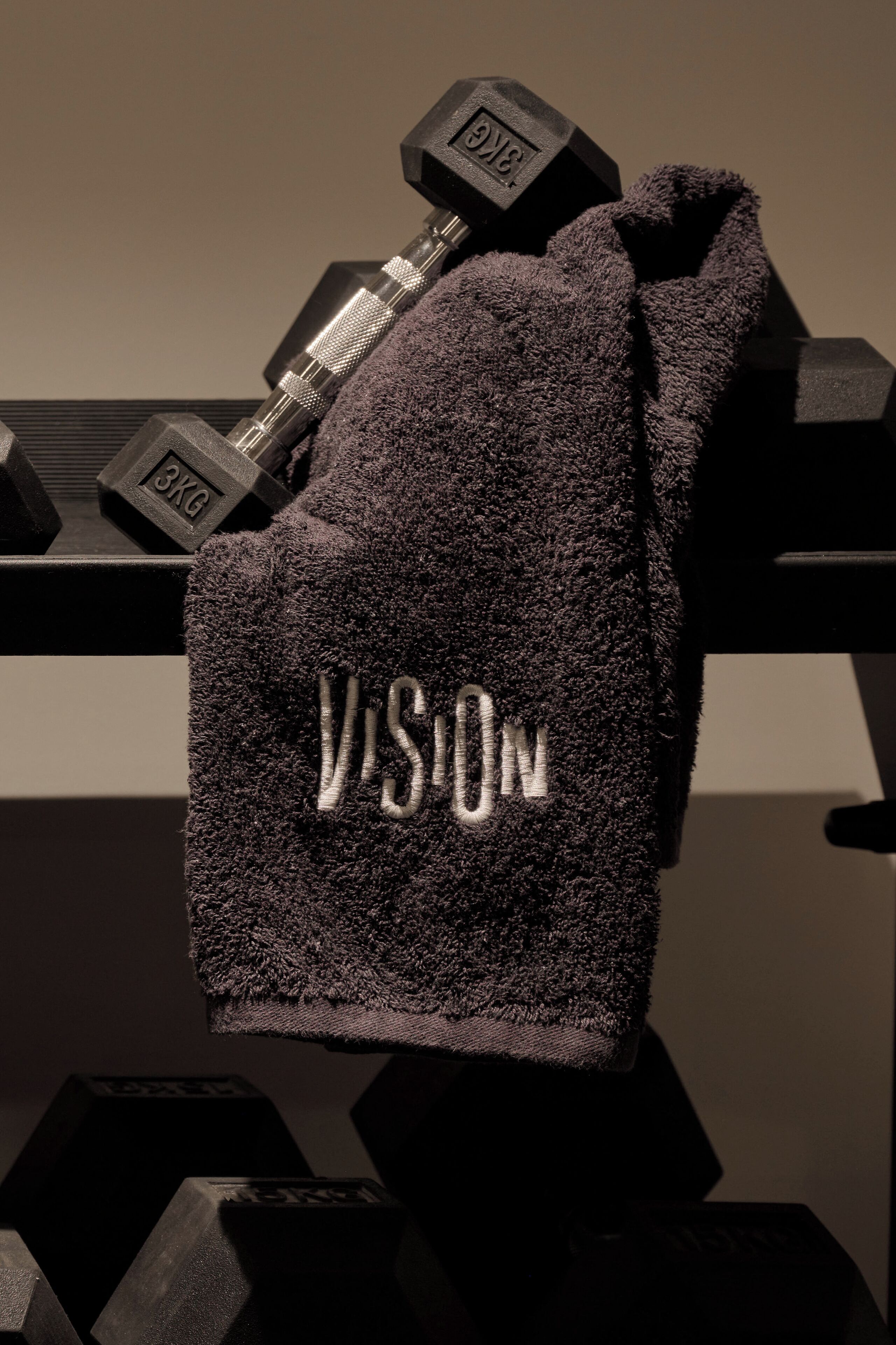 Ein Handtuch mit "VISION"-Stickerei, platziert auf einem Ablagegestell mit Kurzhanteln, die "3KG" beschriften.