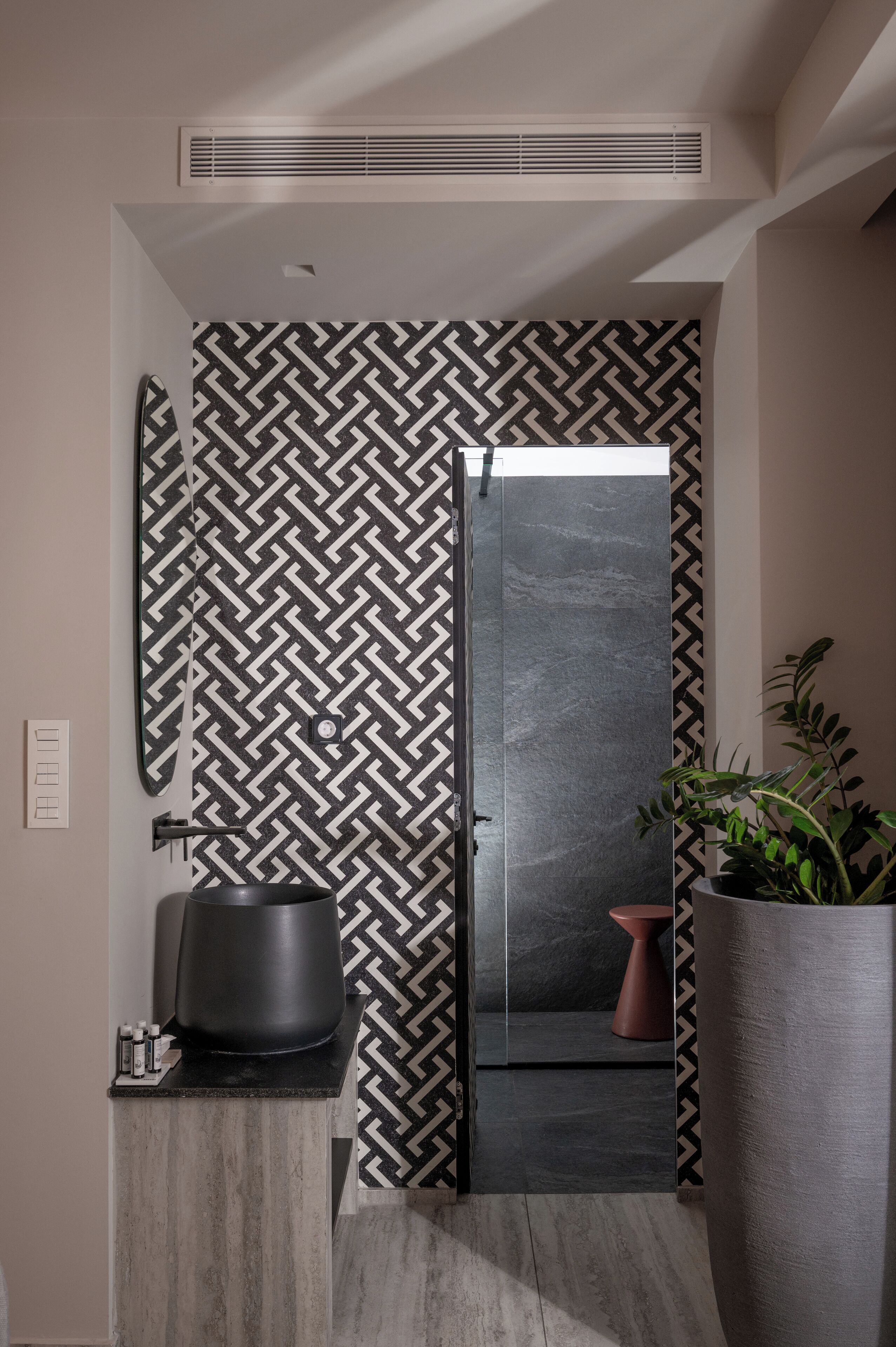 Une salle de bain moderne avec un mur orné d’un motif noir et blanc, un grand miroir, un lavabo noir et une plante en pot.