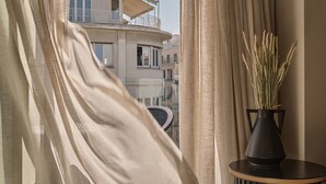 Ein Zimmer mit Blick auf ein Gebäude, durchscheinende Vorhänge, ein Blumentopf auf einem Beistelltisch und ein gemusterter Boden.