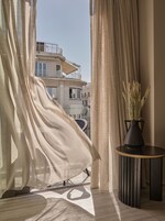 A room with a view of a building through sheer curtains, a potted plant on a side table, and a patterned floor.