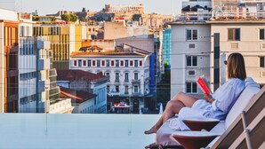 Eine Person entspannt sich auf einem Balkon mit einem Buch und blickt auf eine Stadtansicht mit historischen Gebäuden.