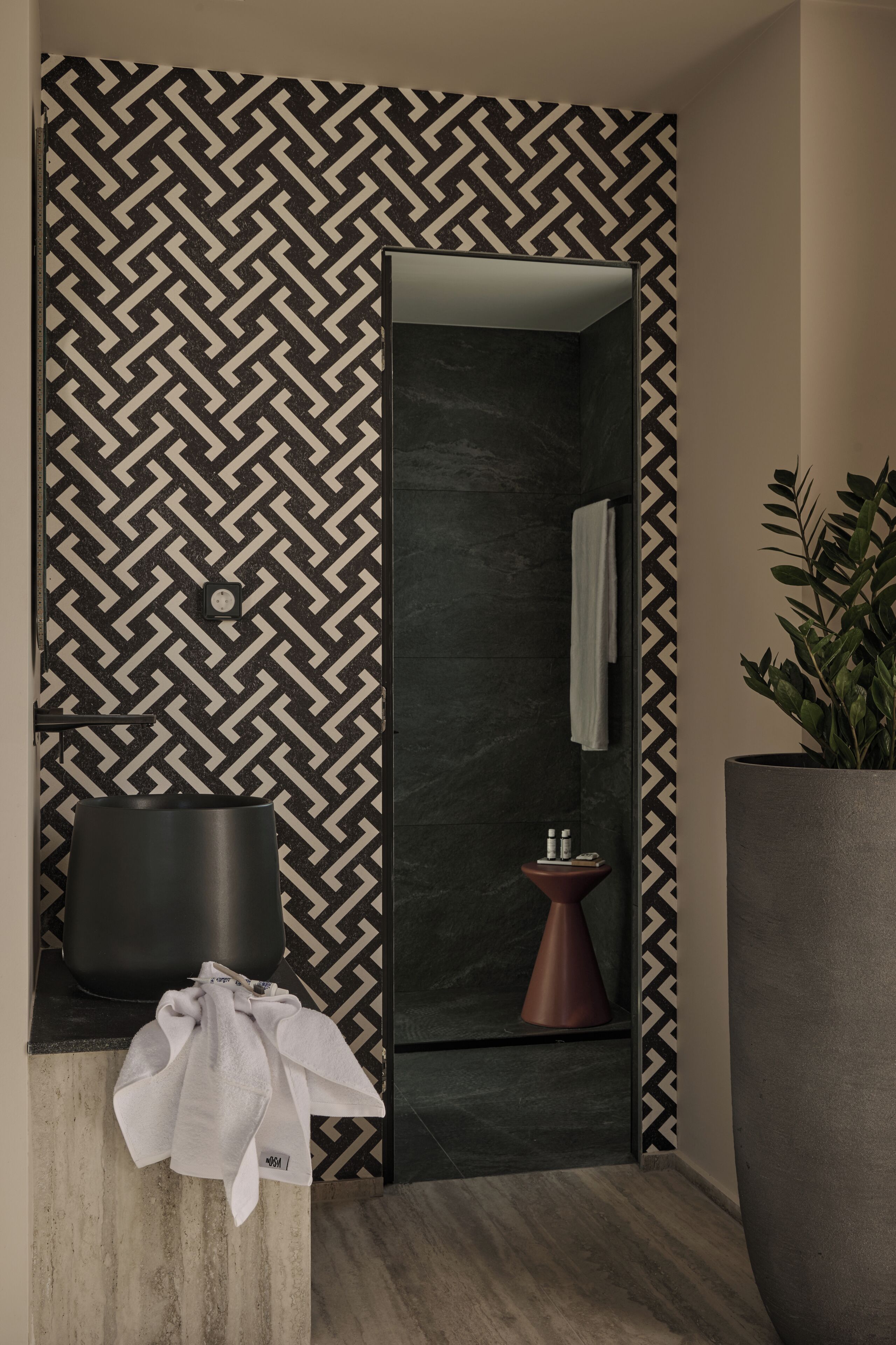 A modern bathroom with a geometric patterned wall, a black sink, a copper basin, and a large potted plant.