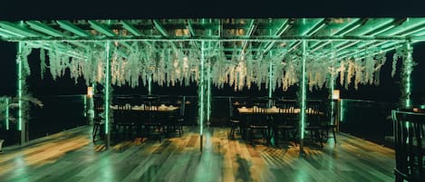 An outdoor seating area with wooden flooring and tables, surrounded by hanging greenery and illuminated by green lights.