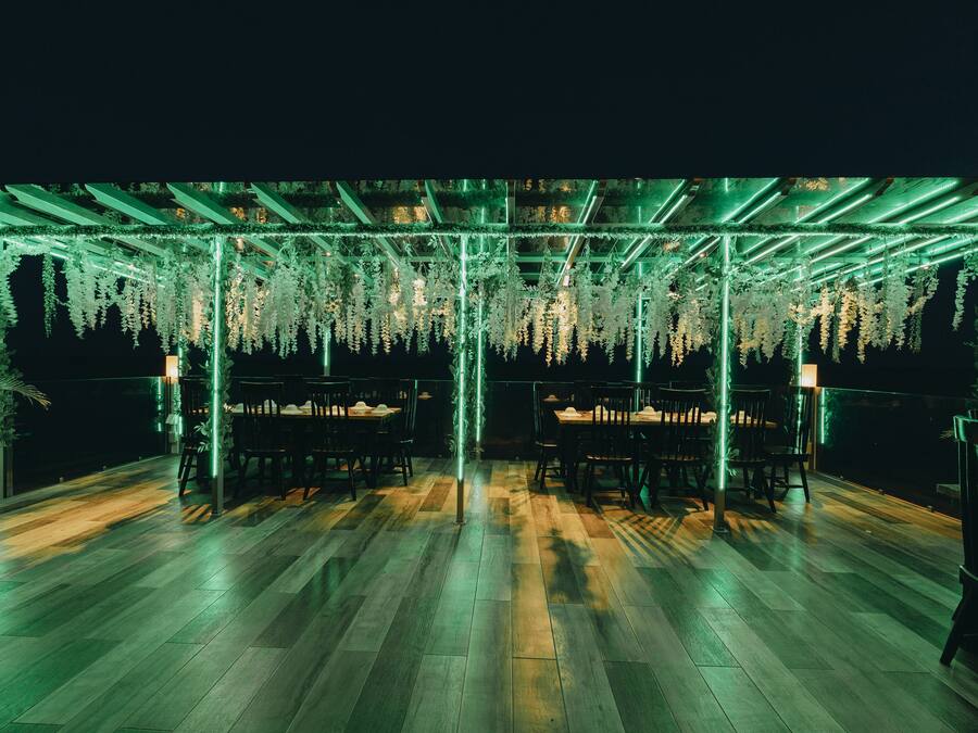 An outdoor seating area with wooden flooring and tables, surrounded by hanging greenery and illuminated by green lights.