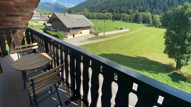 A balcony with a wooden table and bench, overlooking a green field and a mountain.