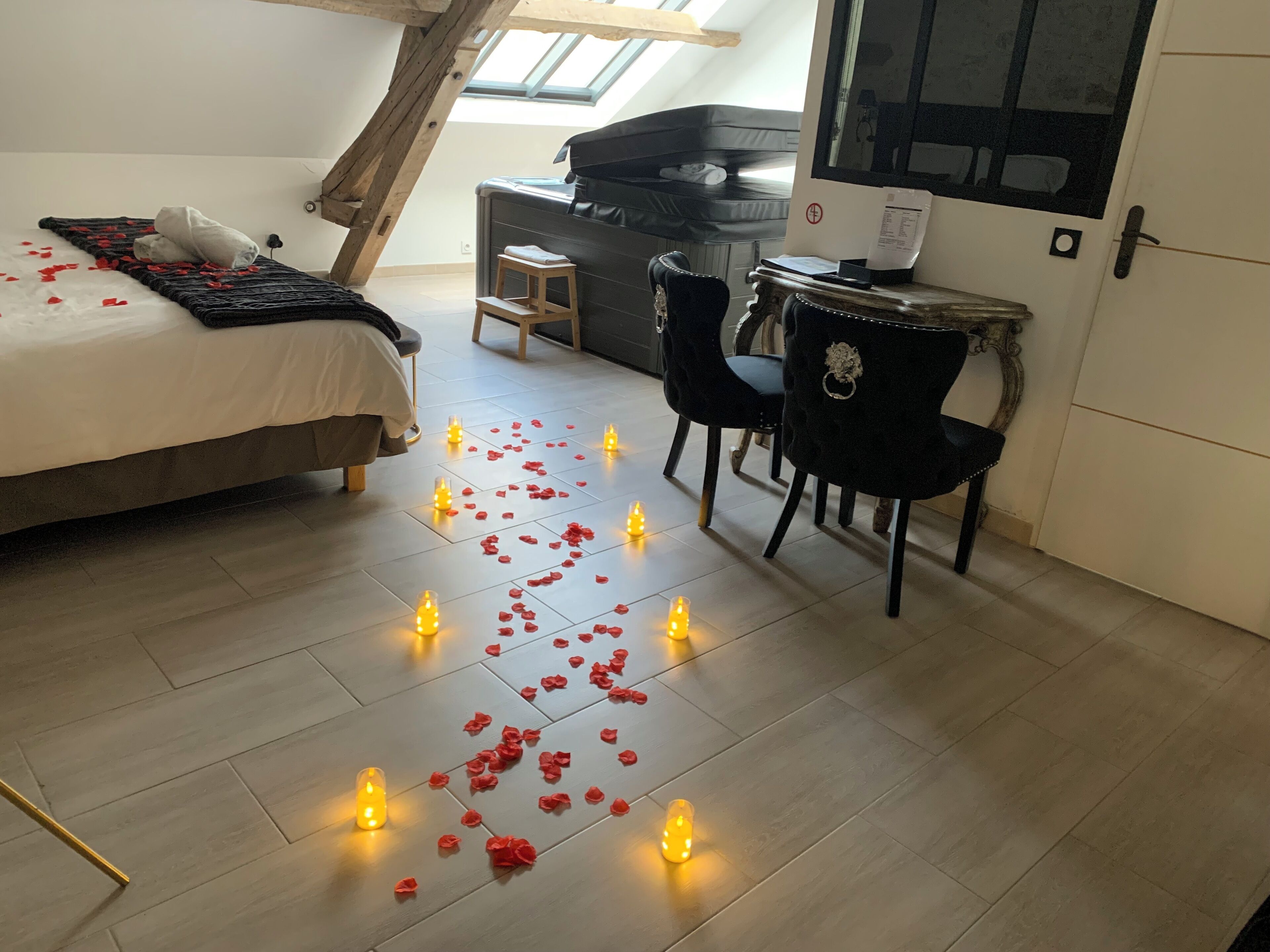 A bedroom with a bed, bedside table, and a small stool. There are candles and rose petals on the floor.