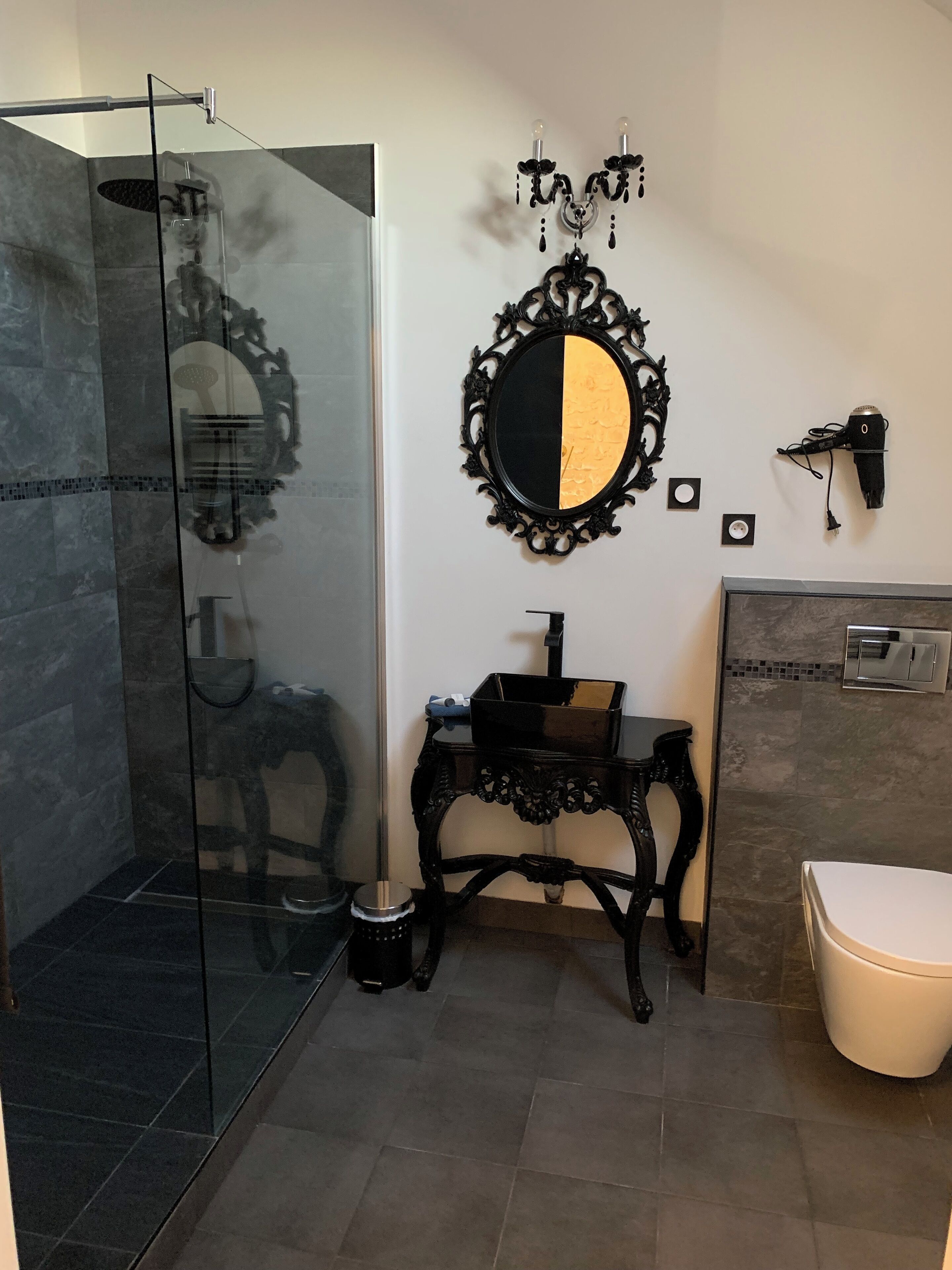 Ein Badezimmer mit Duschkabine aus Glas, einem schwarzen Waschtisch mit Waschbecken, einer Toilette und einem an der Wand befestigten Föhn.