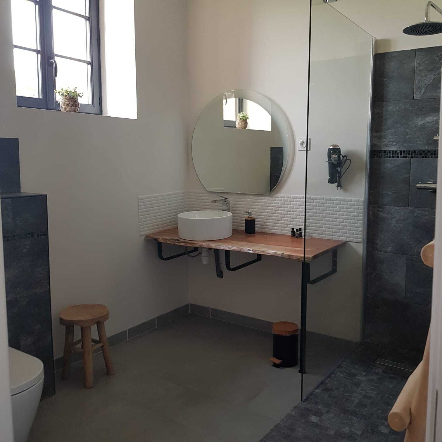 Een moderne badkamer met een ronde spiegel, een witte wastafel, een houten krukje en een douchecabine met glazen wanden.