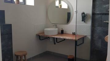 Een moderne badkamer met een ronde spiegel, een witte wastafel, een houten krukje en een douchecabine met glazen wanden.