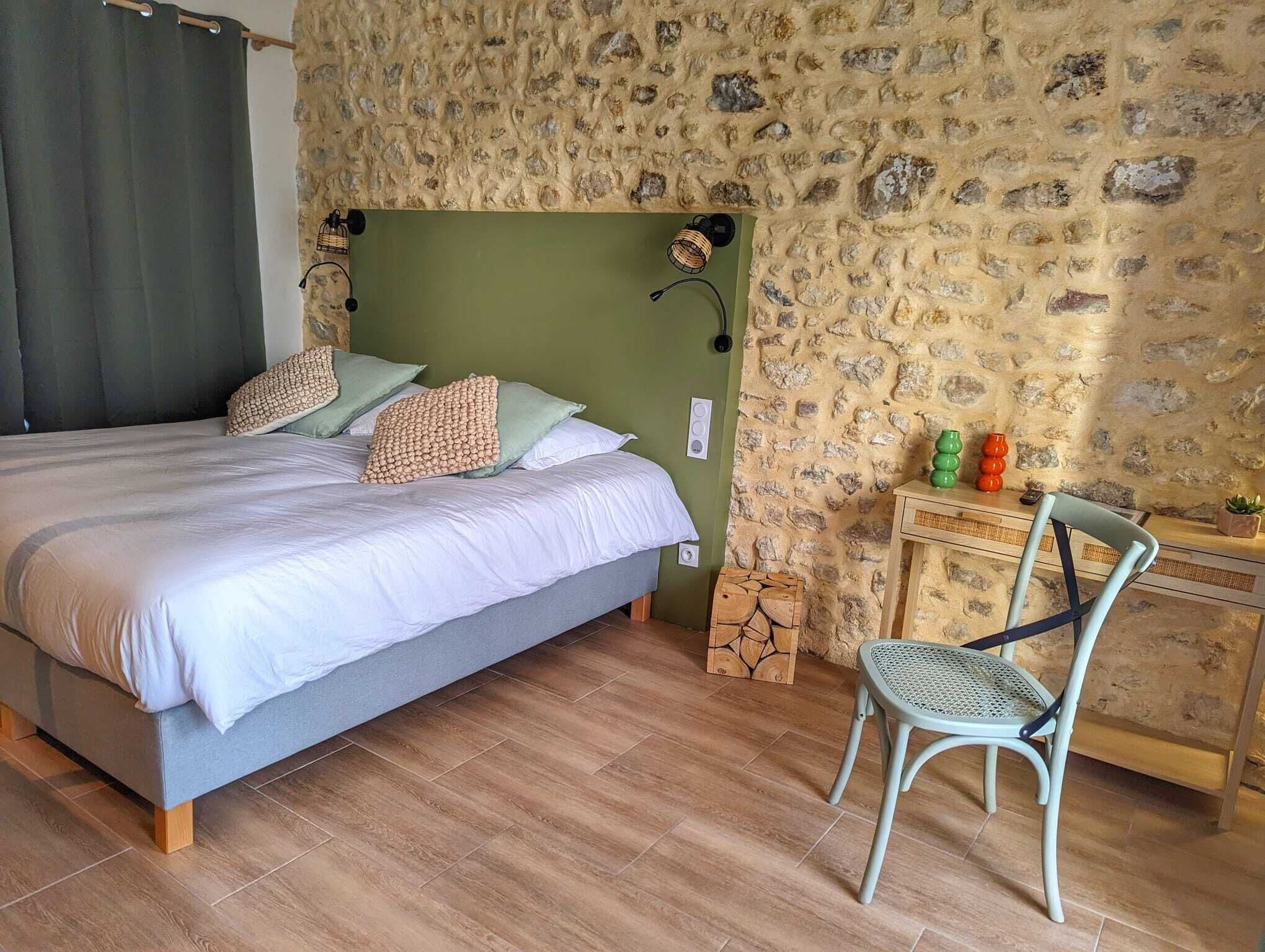Un dormitorio con una cama, una silla, una mesita y una pared de piedra.