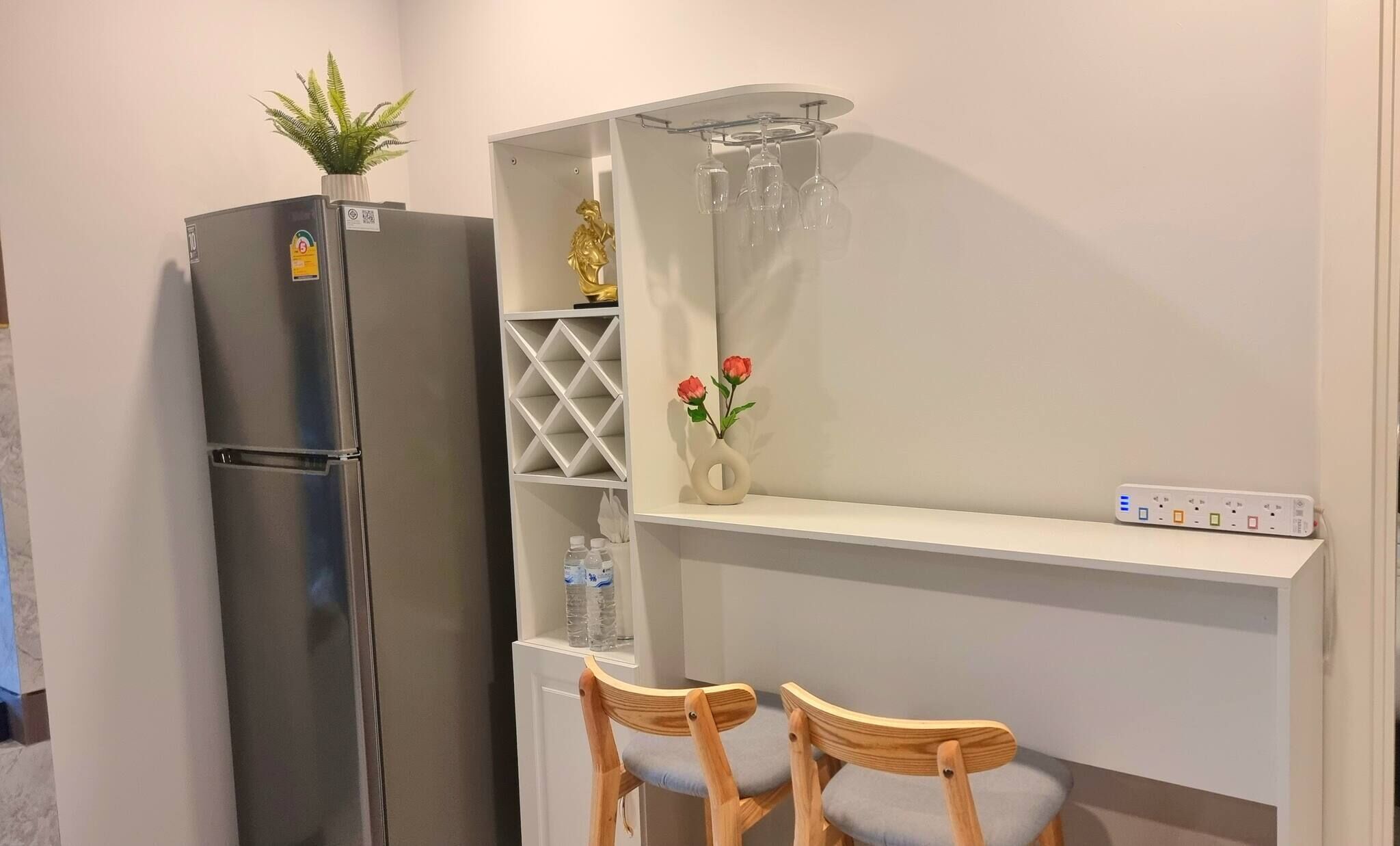 A compact kitchenette with a refrigerator, a small shelf with a vase and a plant, and a bar area with a wine rack and two chairs.