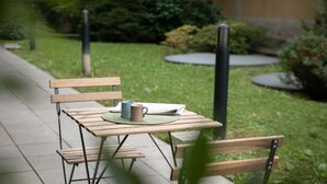 Un banc en bois avec une table et une tasse bleue posée dessus, installé sur un chemin pavé dans un jardin verdoyant, avec un bâtiment en arrière-plan.
