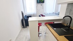 A modern kitchen with a white countertop, a black sink, and a wooden backsplash. There is a TV mounted on the wall, and a window with curtains.