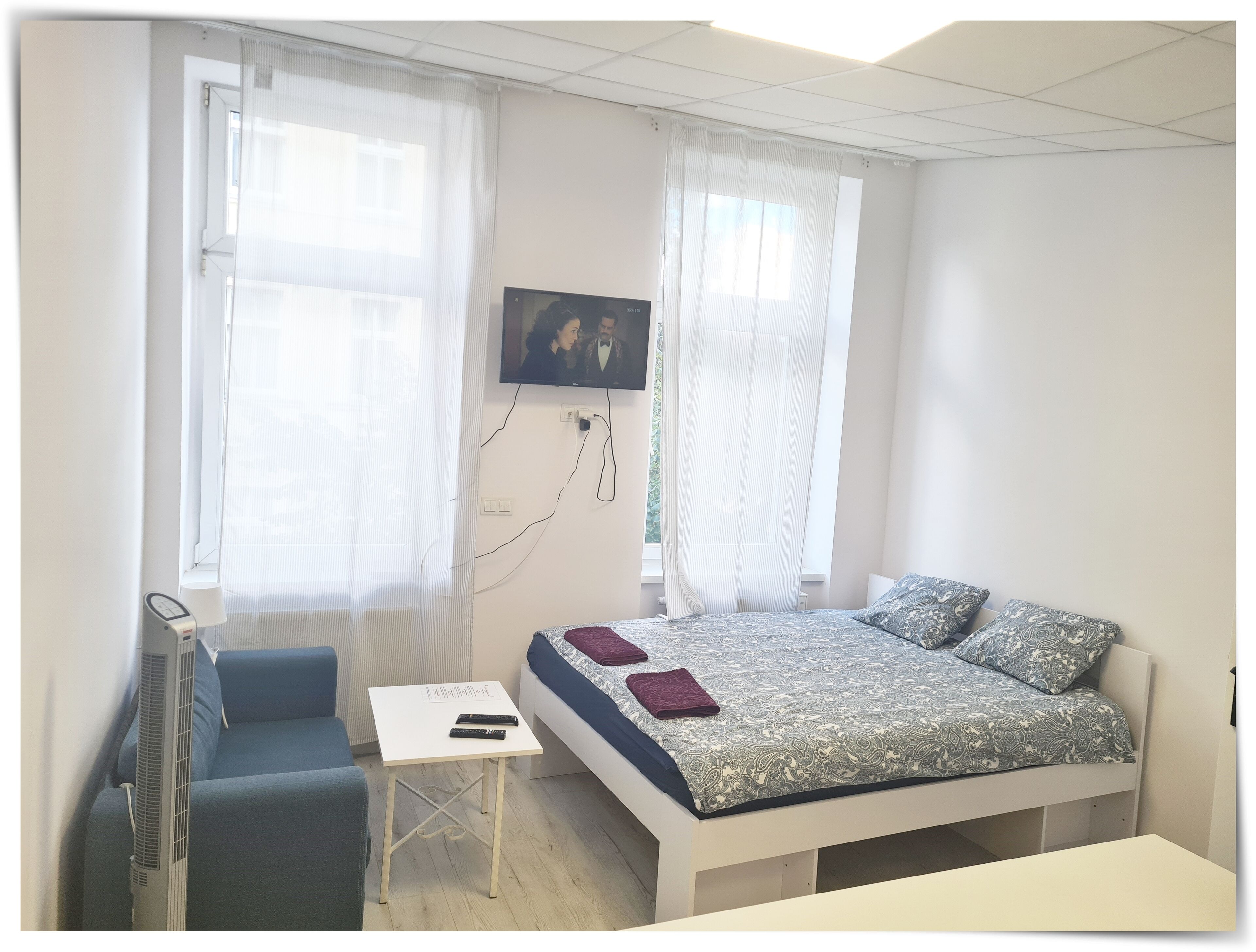 A bedroom with a bed, a desk, a chair, a TV, and a window with curtains.