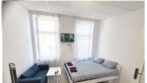 A small, well-lit room with a bed, a sofa, a small table, and a TV mounted on the wall.
