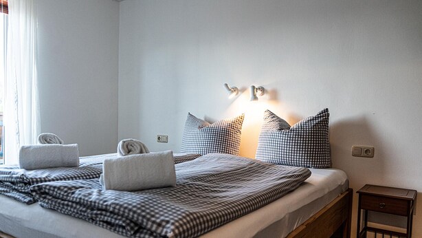 Ein ordentlich bezogenes Bett mit kariertem Bettzeug, zwei zusammengerollte Handtücher und ein Nachttisch mit Lampe.