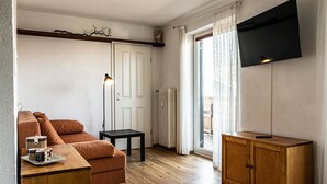 Ein gemütliches Zimmer mit einer Couch, einem kleinen Tisch mit Gläsern, einem Schrank, einer Stehlampe und einem an der Wand befestigten Fernseher.