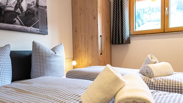 Hotelkamer met twee bedden, geruit beddengoed, een houten garderobe, een lijst met een afbeelding aan de muur en een raam met gordijnen.