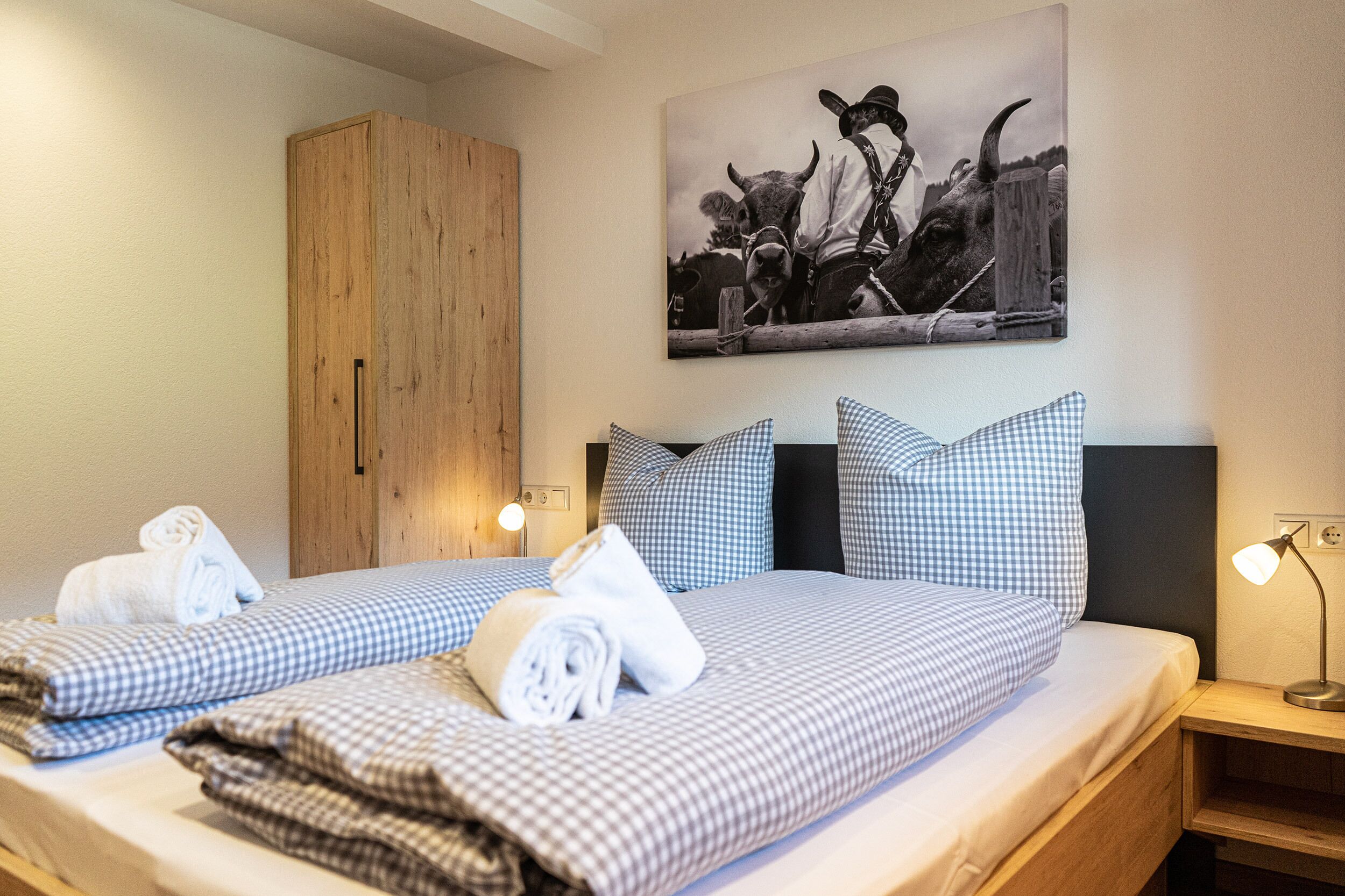 A hotel room with two beds, checkered bedding, a wooden wardrobe, a wall-mounted painting, and bedside lamps.