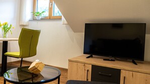 Een moderne woonkamer met een televisie op een houten kast, een groene fauteuil, een glazen tafel en een kleine plant in een pot bij het raam.