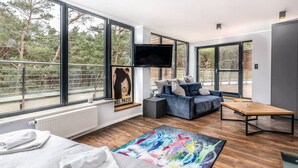 A modern hotel room with a large window, a flat-screen TV, a sofa, a wooden coffee table, and a colorful rug.