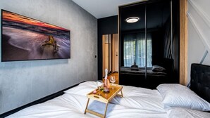 A modern hotel room with a bed, a bedside table with a tray holding a bottle and glasses, a large painting on the wall, and a view of another room through a sliding door.
