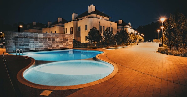 Uma piscina com design curvo, um calçadão pavimentado e um edifício ao fundo.