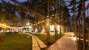 An outdoor patio area with a paved walkway, string lights, and palm trees. - Malnad Shire (Shimoga)