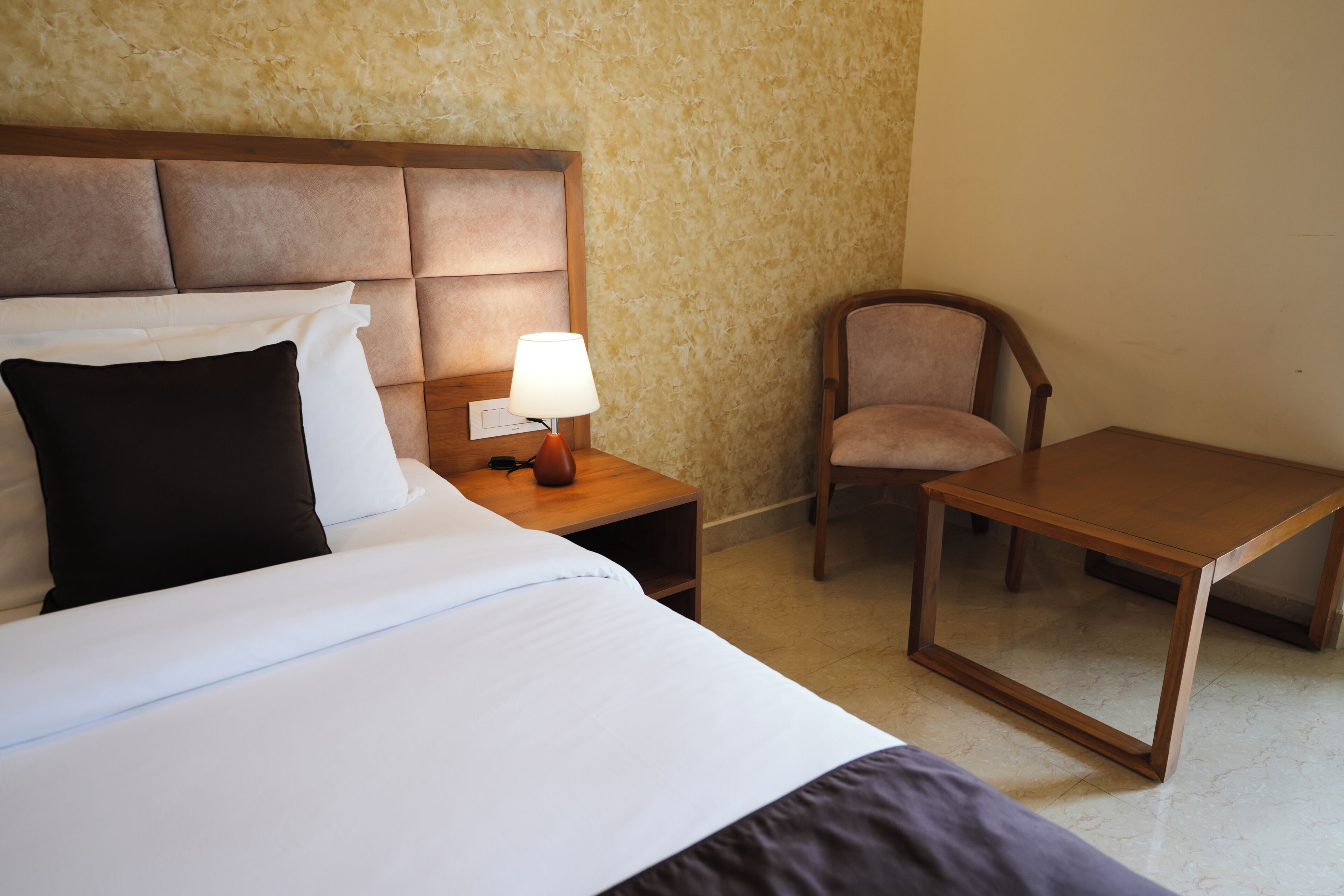 A hotel room with a bed, a nightstand with a lamp, a chair, and a small table.
