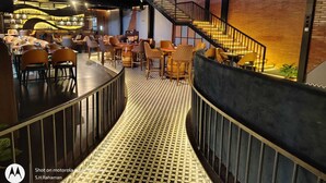 A modern restaurant with a central bar area, multiple seating options, and a staircase leading to an upper level. - Malnad Shire (Shimoga)